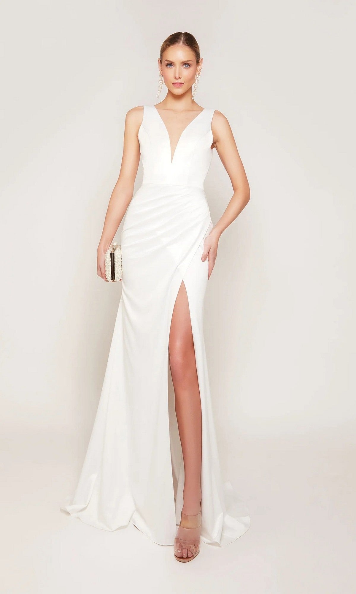 An elegant bride in the Alyce 7092 long formal dress in diamond white, featuring a sleeveless design, deep V-neckline, and high slit, holds a small clutch while standing against a plain light background.